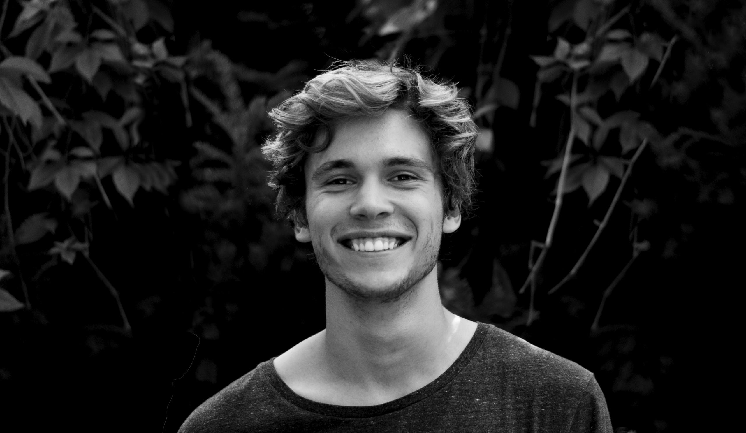A black and white portrait of a young man smiling outdoors with natural background.